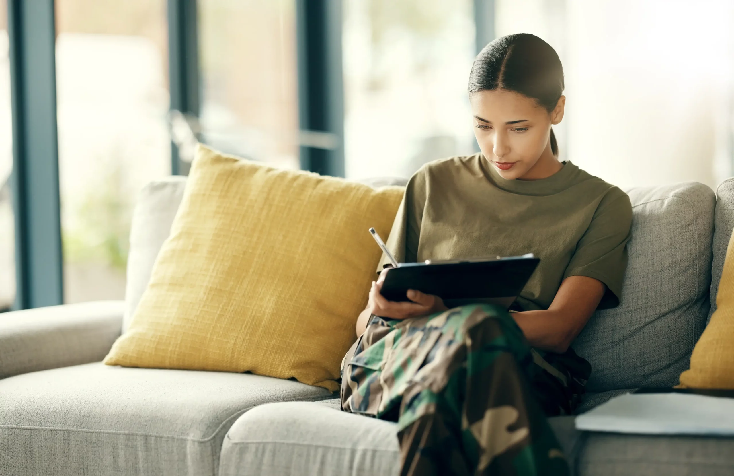 Woman in camo pants filling out paperwork on a couch, preparing for a defence mortgage broker consultation.